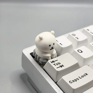 May include: A white keyboard with a small white bear figurine sitting on the Tab key. The bear is sitting on a cloud-shaped base.