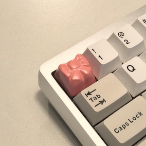 May include: A pink bow-shaped keycap with the word "Tab" printed on the key below it. The keycap is on a white keyboard with black lettering.