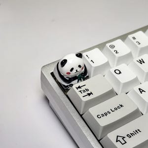 Puede incluir: Un tapón de tecla de panda blanco y negro con un brote de bambú verde en sus patas, sentado en un teclado blanco con teclas negras etiquetadas como "Tab", "Bloq Mayús" y "Shift".