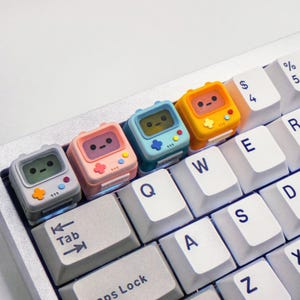 May include: Four colorful keycaps shaped like retro video game consoles with pixelated faces and a joystick design. The keycaps are placed on a white keyboard with black keys.