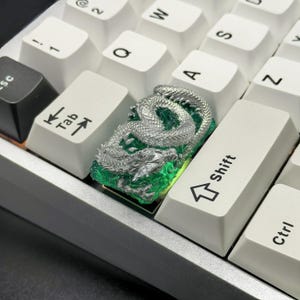 May include: A silver dragon keycap with a green resin base. The keycap is designed to fit a standard mechanical keyboard.