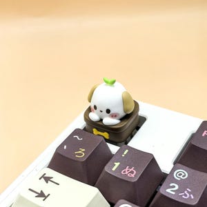 May include: A white and tan puppy-shaped keycap with a green leaf on top, sitting on a brown keycap. The keycap is on a white keyboard with brown keys, some with Japanese characters.