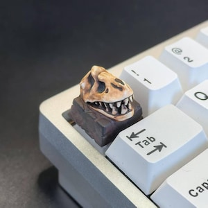 May include: A custom keycap shaped like a dinosaur skull, in shades of brown and tan, sits on a mechanical keyboard. The keycap is detailed with visible teeth and sits on a dark brown base. White keyboard keys are visible.