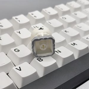May include: A white and gray keycap with a cute cartoon bear design. The keycap is a square shape with rounded corners and a small cross in the center.
