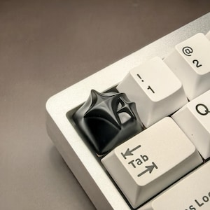 May include: A black keyboard keycap with a unique, abstract design. The keycap is made of a smooth, glossy material and has a slightly raised surface. The keycap is placed on a white keyboard key labelled "Tab" with arrows pointing left and right.