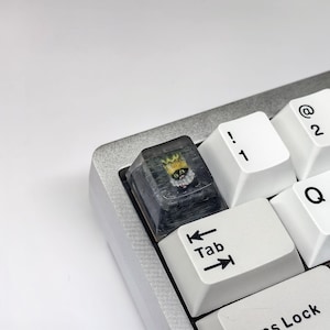 Op de afbeelding: Een aangepaste toetsenborddop met een transparante hars bovenkant en een zwart-wit afbeelding van een kat met een kroon. De toetsenborddop is gelabeld met "Tab" met pijlen die naar links en rechts wijzen.