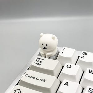 May include: White keyboard keycap with a small white bear figurine sitting on top of it. The keycap says "Tab" and has arrow keys. The bear has black eyes and a small black mouth.