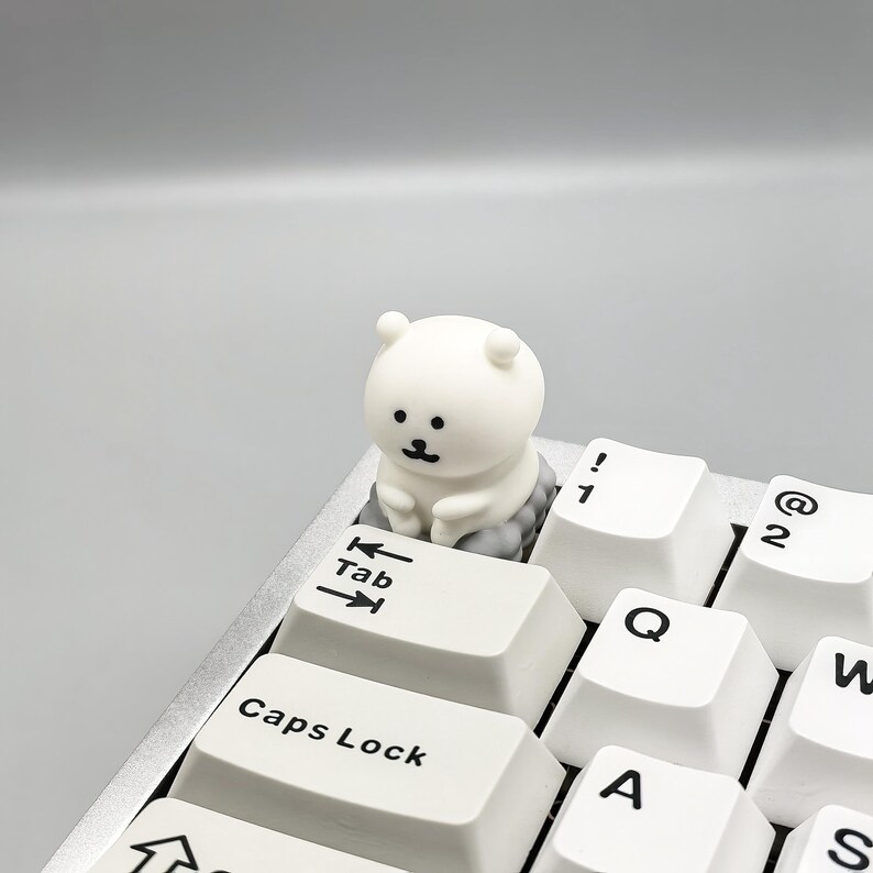 May include: White keyboard keycap with a small white bear figurine sitting on top of it. The keycap says "Tab" and has arrow keys. The bear has black eyes and a small black mouth.