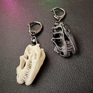 Könnte beinhalten: Zwei Dinosaurierschädel-Schlüsselanhänger. Einer ist cremefarben, der andere schwarz. Beide haben detaillierte Skelettmerkmale und sind an silbernen Schlüsselringen befestigt. Die Schlüsselanhänger werden auf einer dunklen Oberfläche präsentiert.