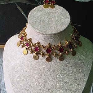 May include: A gold-coloured necklace and matching earrings set. The necklace features red flower-shaped accents and small gold coins. The earrings are small, round, and feature a red flower design. The jewellery is displayed on a mannequin.