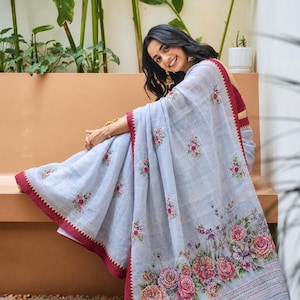 May include: A light blue saree featuring a deep red border with a zigzag edge. The fabric is adorned with small embroidered floral motifs scattered across the body and a large, detailed floral print along the bottom hem in shades of pink and green.