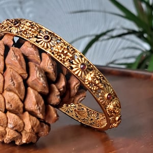 May include: A gold-toned metal bangle bracelet featuring an intricate openwork design of repeating floral motifs and small, circular dark red stones embedded at regular intervals along the outer surface of the circular band.