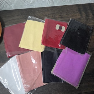 Puede incluir: Una colección de artículos cuadrados de tela de colores, cada uno empaquetado individualmente en plástico transparente. Los colores incluyen blanco, rosa, negro, morado, rojo, amarillo y burdeos. Los artículos están dispuestos sobre una superficie de madera oscura.