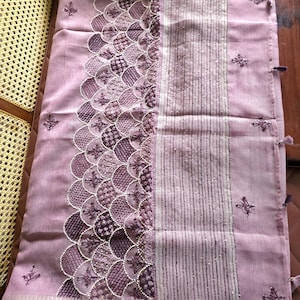 May include: A lilac textile with detailed embroidery and embellishments. The design includes a scalloped pattern on one side and vertical stripes on the other. The edges are trimmed with a decorative border and tassels.