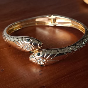 Pode incluir: Uma pulseira de cobra dourada com duas cabeças de cobra voltadas uma para a outra. Cada cabeça de cobra tem um olho de gema verde e pequenas pedras preciosas transparentes. A pulseira está em uma superfície de madeira.