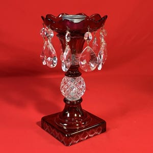May include: A ruby red glass candle holder with clear crystal teardrop pendants. The candle holder has a square base, a diamond-cut sphere, and a scalloped top. The background is a solid red.