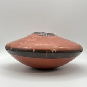 May include: A terracotta-colored ceramic vase with a wide, flattened shape. The vase features a dark brown band around the middle and a lighter, etched design on the upper portion. The overall design is smooth and modern.