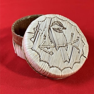 May include: A round, lidded box with a woven design. The lid features a detailed pattern of white sticks arranged in a geometric design. The box is open, revealing a brown interior and a red background.