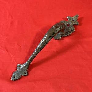 May include: A dark bronze-toned metal door handle featuring a curved, textured grip and two ornate, decorative mounting plates at each end. Each mounting plate includes two small circular screw holes for attachment to a door surface.