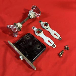 Puede incluir: Juego de herrajes de puerta antiguos, que incluye dos pomos de puerta de cristal transparente, una cerradura de embutir de metal negro, dos placas traseras pintadas de blanco y tornillos. Fondo rojo.