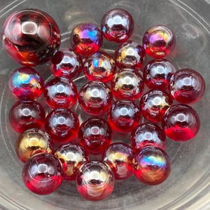 Può includere: Una collezione di biglie di vetro rotonde di varie dimensioni, principalmente di colore rosso intenso, alcune con riflessi iridescenti simili all'arcobaleno e punti luce riflettenti, il tutto contenuto in una ciotola di vetro trasparente.