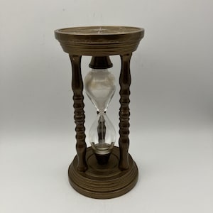 May include: A bronze-colored hourglass with a clear glass center filled with white sand. The hourglass has a decorative, ornate design with a round base and top, and four supporting pillars. The top has a star-like design.