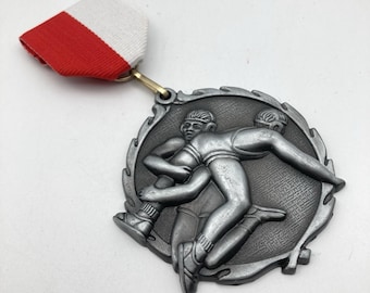 May include: A silver-toned circular medal featuring a relief sculpture of two wrestlers in a grappling hold. The medal is attached to a folded ribbon with a horizontal red stripe on the bottom and a white stripe on the top, connected by a gold ring.