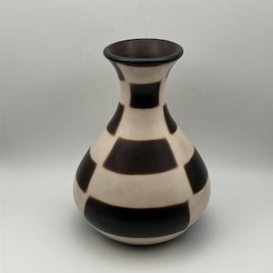 Puede incluir: Un jarrón decorativo con un cuerpo bulboso y un cuello estrecho. El jarrón presenta un fondo color crema con patrones cuadrados de color marrón oscuro. El borde y el interior del jarrón son de color marrón oscuro.