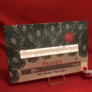 Può includere: Una scatola di 100 luci in miniatura Ultra Bright di Frank. La scatola ha un design con aghi di pino e pigne. Il testo "Ultra Bright" e "Where Beautiful Things Begin" sono visibili. La catena di luci è lunga 3,81 metri.