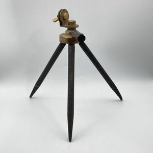 May include: A vintage tabletop tripod featuring three slender, dark-toned metal legs and a brass-colored mounting head. The head includes an adjustable circular dial with a small wing nut and a black plastic handle for positioning the camera mount.