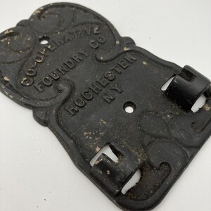 以下が含まれることがあります： CO-OPERATIVE FOUNDRY CO. ROCHESTER NYと浮き彫り文字が刻まれた黒い鋳鉄製のストーブプレート。長方形の金属パーツには装飾的なスクロール模様、2つの取り付け穴、および2つの統合されたヒンジブラケットが施されています。