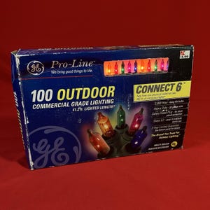 Pode incluir: Uma caixa de luzes de Natal GE Pro-Line 100 Outdoor. A caixa é azul com o texto "100 Outdoor" e "41.2 ft. Lighted Length", que são aproximadamente 12,56 metros. A caixa também tem uma sequência de luzes coloridas.