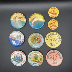 May include: A collection of vintage round pinback buttons with various designs and text. Some buttons feature text like "Tip-Up Town U.S.A." and "Mid-Forest Lodge." Colors include blue, yellow, red, and green.