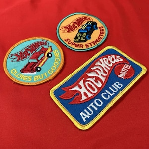 May include: Three embroidered patches with the Hot Wheels logo and car designs. One patch is rectangular with "Auto Club" text, another is round with "Super Streeters", and the third is round with "Oldies But Goodies". The patches have a variety of colors including red, blue, yellow, and orange.
