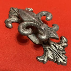 May include: A silver-colored, fleur-de-lis shaped cabinet pull. The decorative hardware features a detailed design with curved lines and a textured finish. The pull is set against a red background.