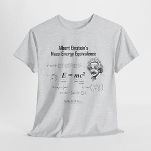 Pode incluir: Uma camiseta cinza com uma ilustração em preto e branco de Albert Einstein e a famosa equação E=mc². O texto "Albert Einstein's Mass-Energy Equivalence" é impresso acima da ilustração. A camiseta apresenta várias equações de física relacionadas à equivalência massa-energia.