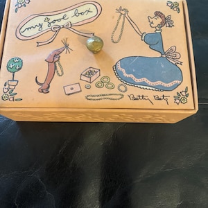 May include: A vintage, light brown, rectangular jewelry box with a cartoon illustration of a woman and a dog. The box has a gold knob and the text "my jewel box" on the lid. The text "Betty Bett" is signed on the bottom right corner of the box.