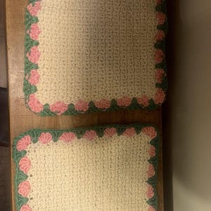 May include: Two off-white, rectangular crochet placemats with a scalloped border. The border features alternating pink flower shapes and green leaves. The placemats are placed on a wooden surface.