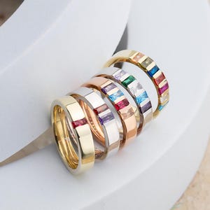 May include: Five gold bands with different colored gemstones set in a row. The gemstones are red, purple, blue, green, and yellow.