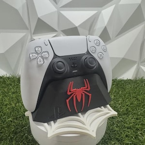 May include: A white and black video game controller resting on a black charging dock. The dock features a prominent red spider emblem on the front and sits atop a white, web-shaped base. The controller has standard buttons and joysticks.