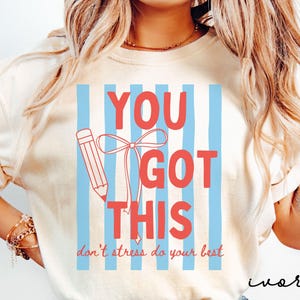 May include: Ivory-colored t-shirt with the text "YOU GOT THIS" in red, set against a backdrop of blue vertical stripes. A red pencil with a bow is also featured. The phrase "don't stress do your best" is written below.