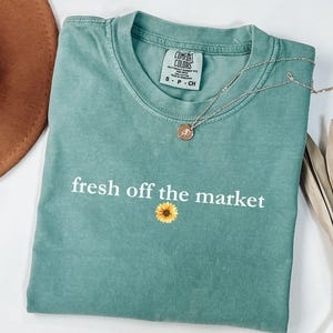 May include: A teal blue t-shirt with the text "fresh off the market" in white lettering and a yellow sunflower graphic. The shirt is made of a soft, comfortable fabric.