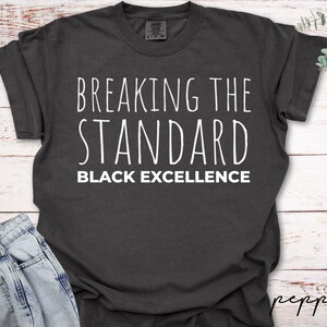 May include: Dark gray t-shirt with the words "BREAKING THE STANDARD BLACK EXCELLENCE" in white lettering. The shirt is slightly wrinkled and appears to be made of a soft material. The shirt is displayed on a white wooden surface.
