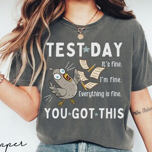May include: Dark gray t-shirt with the text "TEST DAY" and a cartoon bird with flying papers. The shirt also says "It's fine. I'm fine. Everything is fine. YOU GOT THIS."