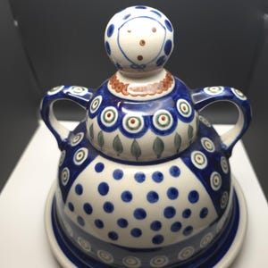 Puede incluir: Una mantequera de cerámica con tapa, con un diseño caprichoso. El plato es principalmente blanco con lunares azules y bandas azules decorativas. La tapa está rematada con una figura estilizada con una cara pintada.