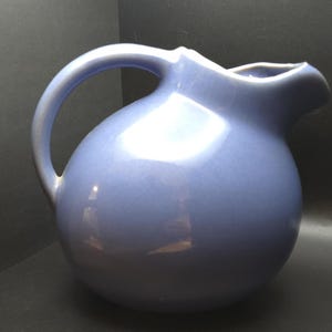 Op de afbeelding: Een lichtblauwe keramische kan met een afgeronde body en een gebogen handvat. De kan heeft een tuit om uit te schenken en een glanzende afwerking. De kan is een klassiek ontwerp, geschikt voor het serveren van dranken.