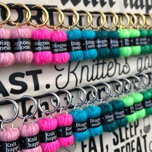 Könnte beinhalten: Nahaufnahme einer weißen Stofftasche mit dem aufgedruckten Text "Life is Knit happens!" Die Tasche ist mit Reihen von bunten Garn-Schlüsselanhängern verziert, die jeweils einen kleinen Metallring haben. Die Schlüsselanhänger sind in einem Muster aus verschiedenen Farben angeordnet, darunter Pink, Lila, Blau, Grün und Braun.