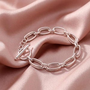 May include: A silver-colored chain bracelet with an oval link design. The bracelet features a toggle clasp closure. The jewelry is displayed on a pink satin fabric background.