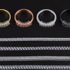 Puede incluir: Cuatro anillos en oro, cobre, plata y negro, cada uno con un ajustador de anillo de plástico transparente. Debajo, se muestran varios medidores de anillos de plástico transparente, que ofrecen una variedad de tamaños. Ideal para joyería.