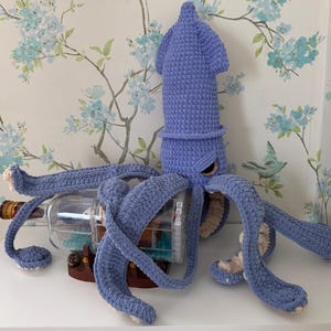 May include: A light purple crocheted squid plush toy with a textured surface, featuring long tentacles with cream-colored underside details. The squid is positioned around a clear glass bottle containing a miniature ship model on a brown base.